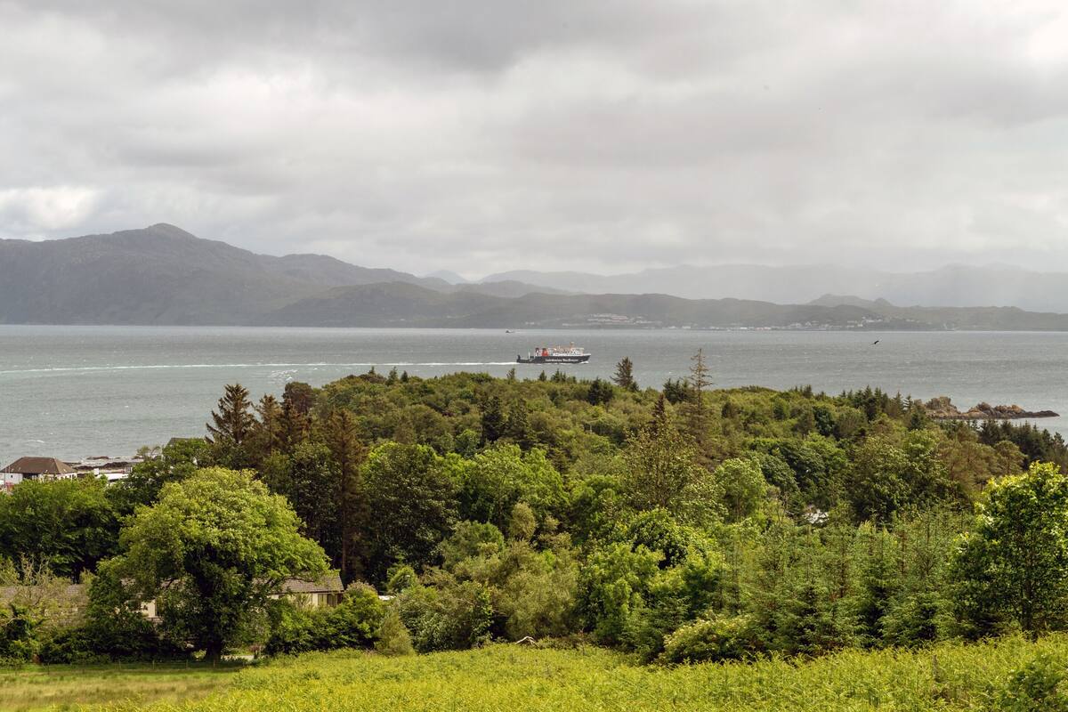 A coastal landscape with a boat on the water, dense greenery, and distant mountains.