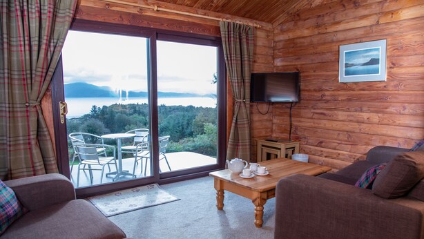 Een gezellig vakantiehuisje met houten inrichting, een bank, een tafel met theepotten, en uitzicht op een meer en bergen door grote glazen deuren.
