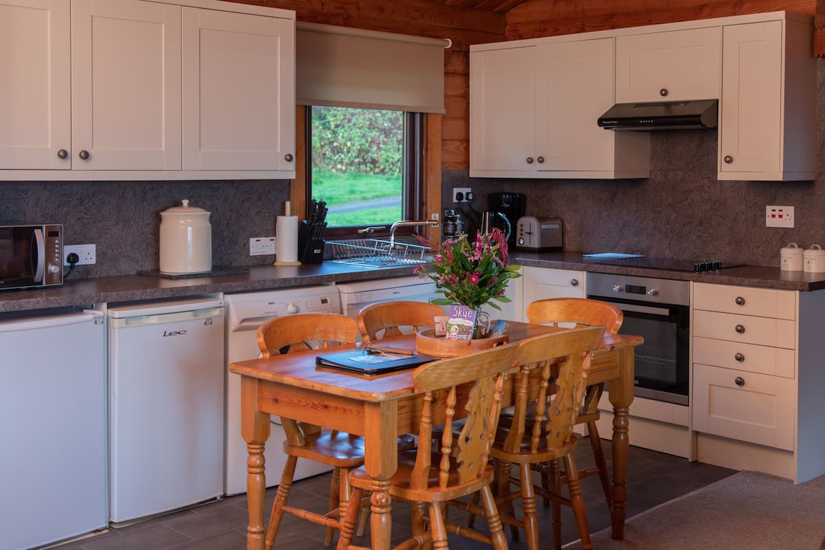A modern kitchen with white cabinets, a dining table with chairs, a coffee maker, and a view of greenery outside.