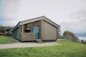 A single-story cabin with a sloped roof, a front porch with a bench, and a view of a lake. - Armadale Castle Cabins (Sleat)