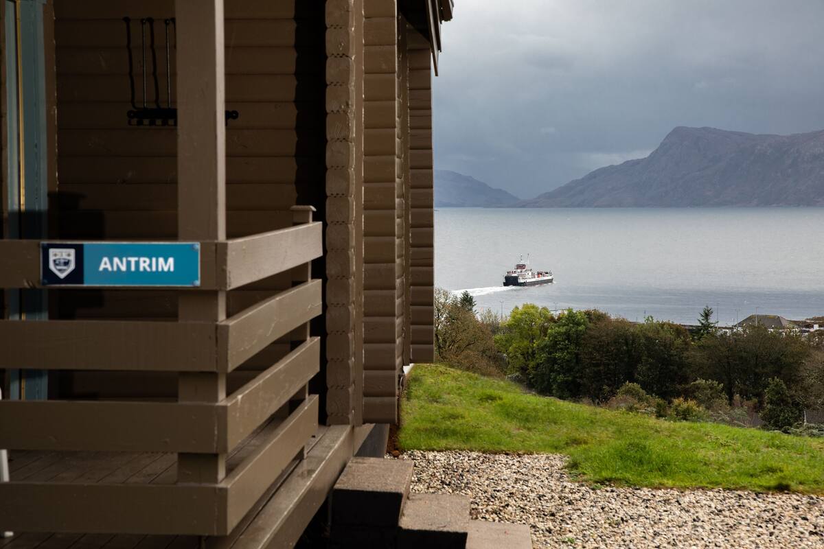 A view from a building with a sign reading "ANTRIM" overlooking a calm body of water with a boat in the distance and mountains in the background.