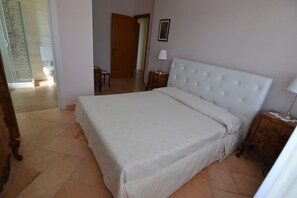 Un dormitorio con una cama grande, una mesita de noche de madera, una lámpara blanca y una puerta que da a otra habitación.