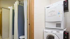A laundry area with a washing machine and dryer, a small stool, and a shower curtain. - Albergue Albas - Hostel (Logroño)