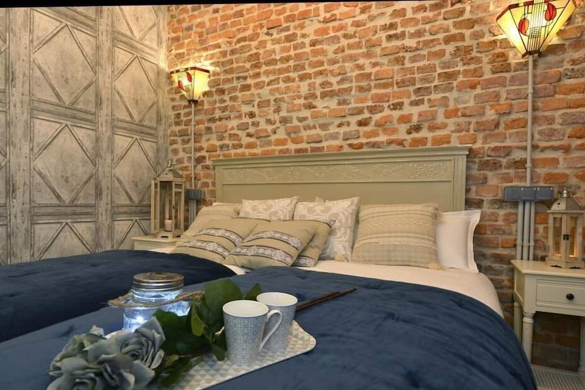 A bedroom with a brick wall, a wooden door, a bed with blue bedding, two bedside tables with lamps, and a tray with a candle, a vase, and two cups.