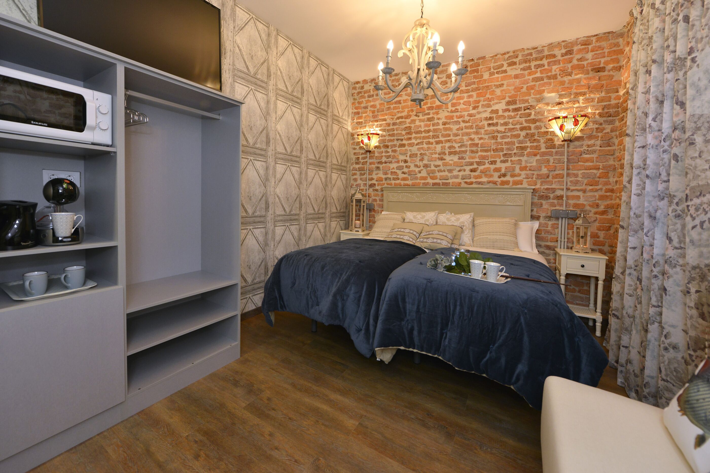 A bedroom with a brick wall, a bed with a blue bedspread, a white wardrobe, and a chandelier.