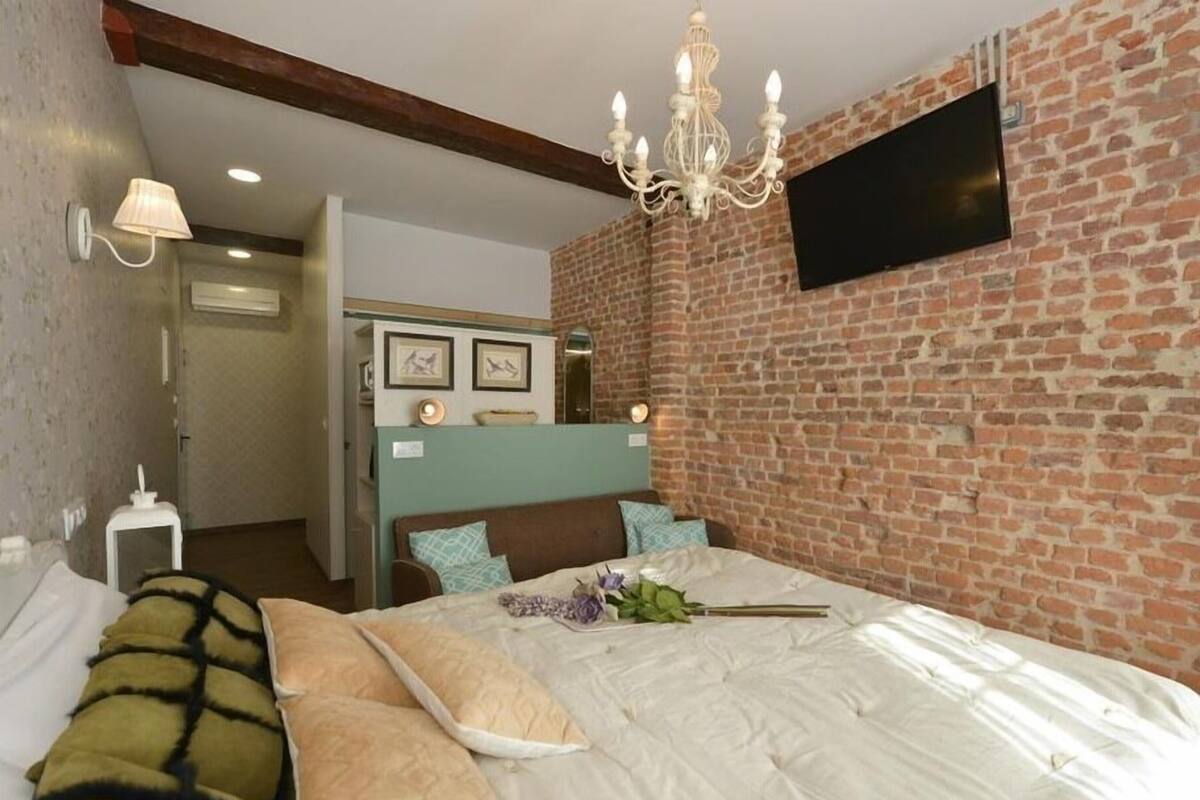 A bedroom with a brick wall, a bed with multiple pillows, a chandelier, and a flat-screen TV mounted on the wall.