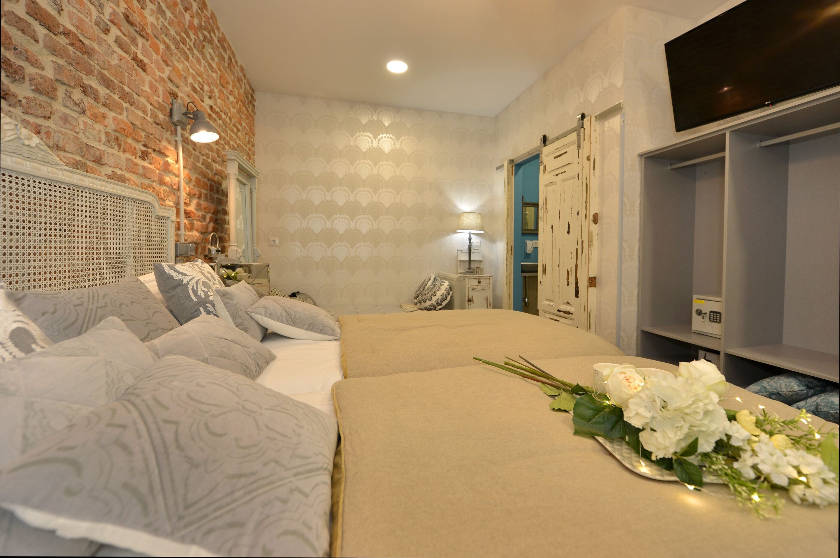 Un dormitorio con pared de ladrillo, cama con ropa de cama estampada, mesita de noche con lámpara y televisor en la pared.