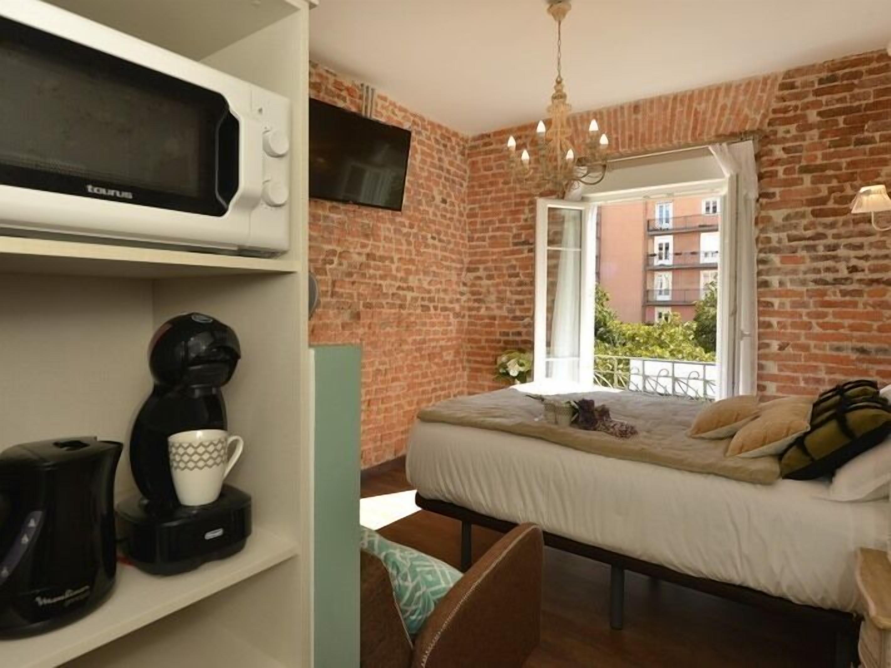 A bedroom with a brick wall, a bed, a TV, a coffee maker, and a window with a balcony view.