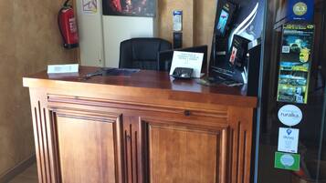 A reception desk with a wooden finish, a black office chair, and a computer monitor. There are various signs and posters on the wall, including one for a fire extinguisher and another for a recommended local attraction.