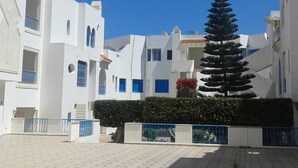 A courtyard with white buildings, a tall conifer tree, and a patterned floor. - El Goulli Appartement (Hergla)