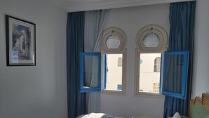 A bedroom with a bed, two windows with blue frames, a painting on the wall, and a lamp on the bedside table. - El Goulli Appartement (Hergla)