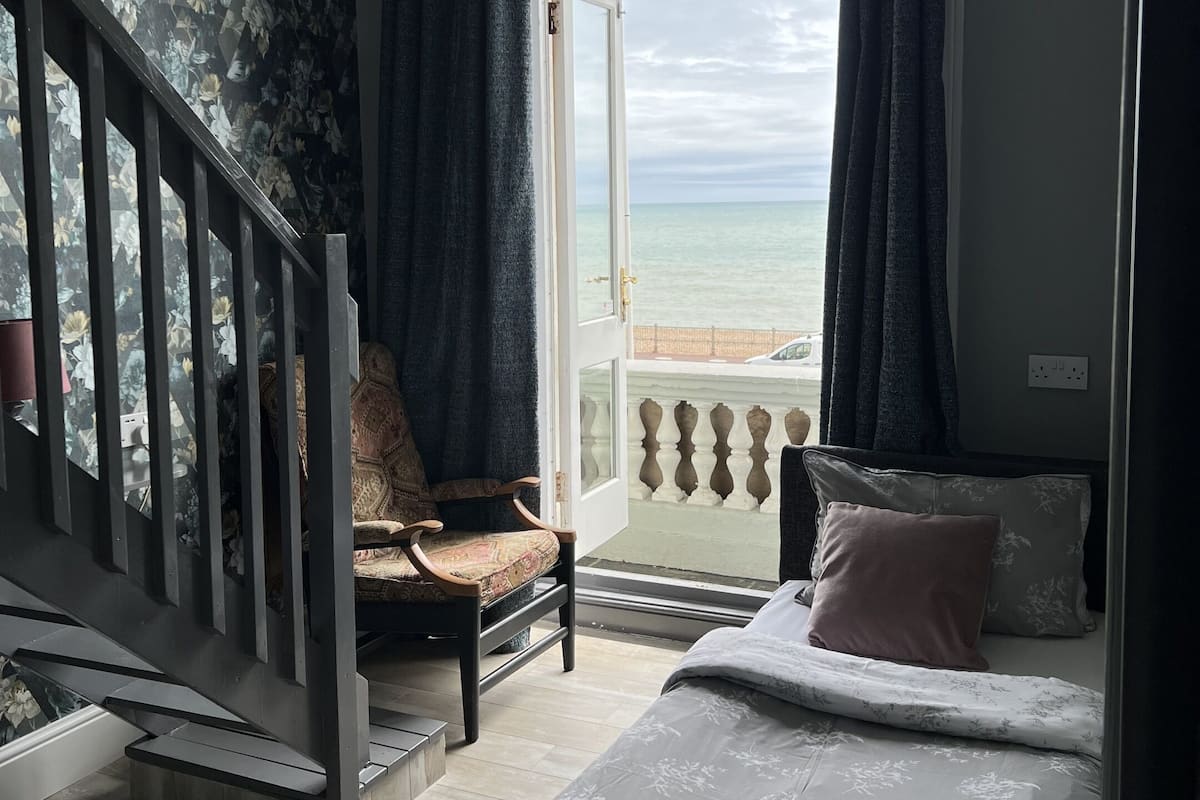 A bedroom with a bed, a chair, a staircase, and a view of the sea through an open door.
