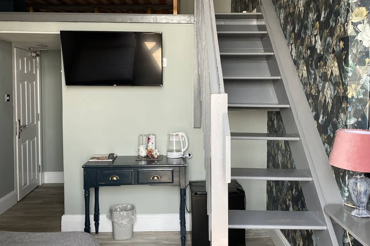 A modern bedroom with a loft bed, a TV mounted on the wall, a desk with a coffee maker, and a staircase with floral wallpaper.