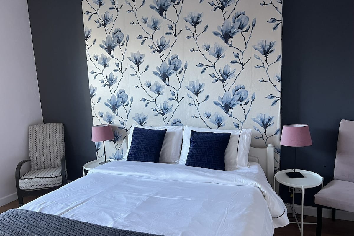 A neatly made bed with white and blue bedding, a chair, a small bedside table with a lamp, and a wall with a floral pattern.