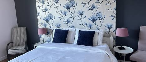 Una cama bien hecha con ropa de cama blanca y azul, una silla, una mesita de noche con una lámpara, y una pared con un estampado floral.