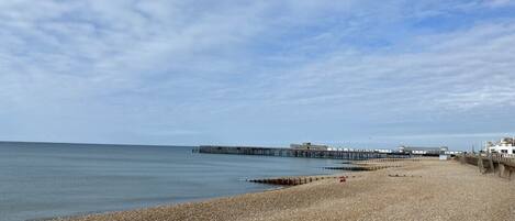 A pebbled beach with a pier extending into the sea, a clear sky, and a building in the background.