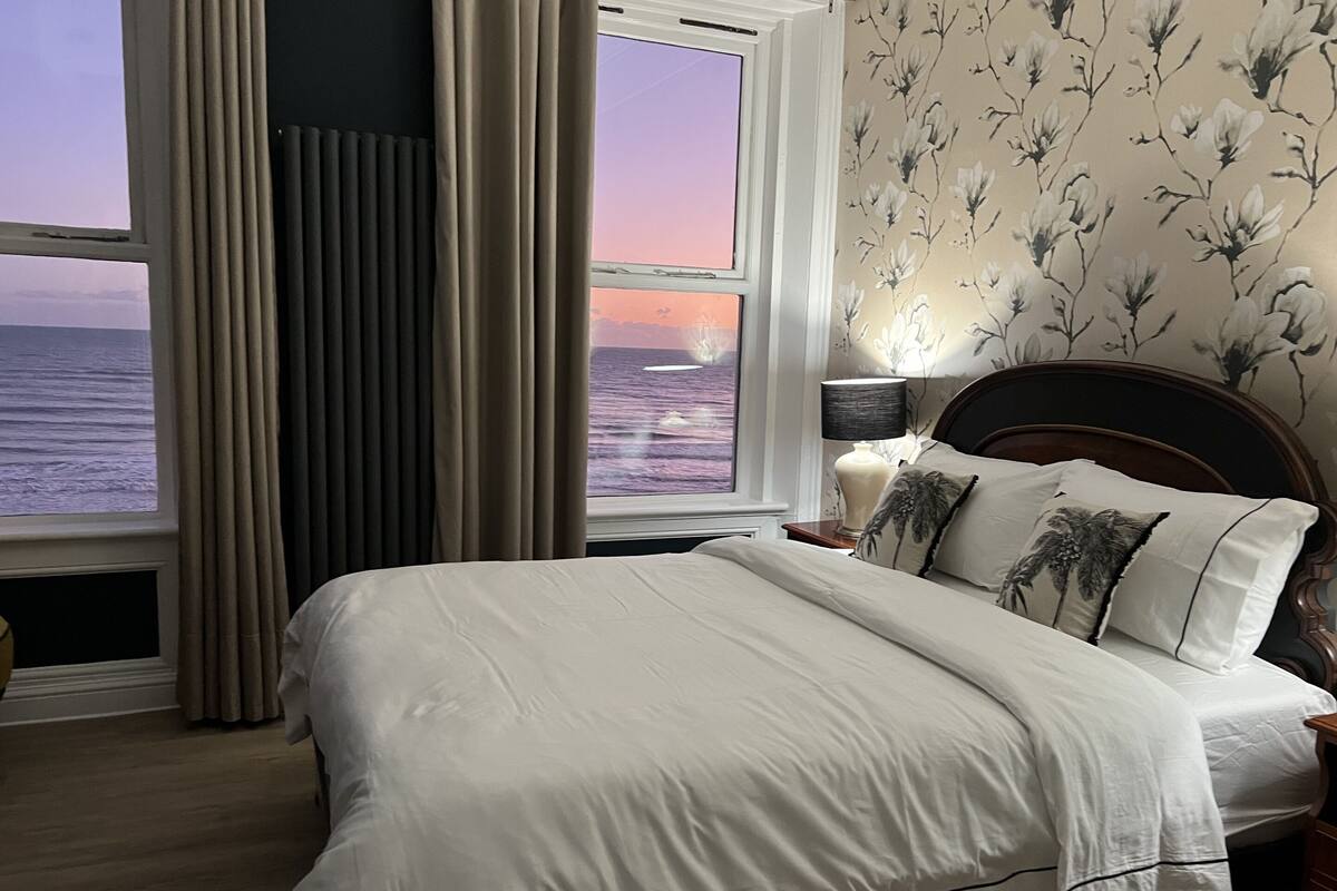 A bedroom with a large bed, a nightstand with a lamp, and a view of the ocean through a window.