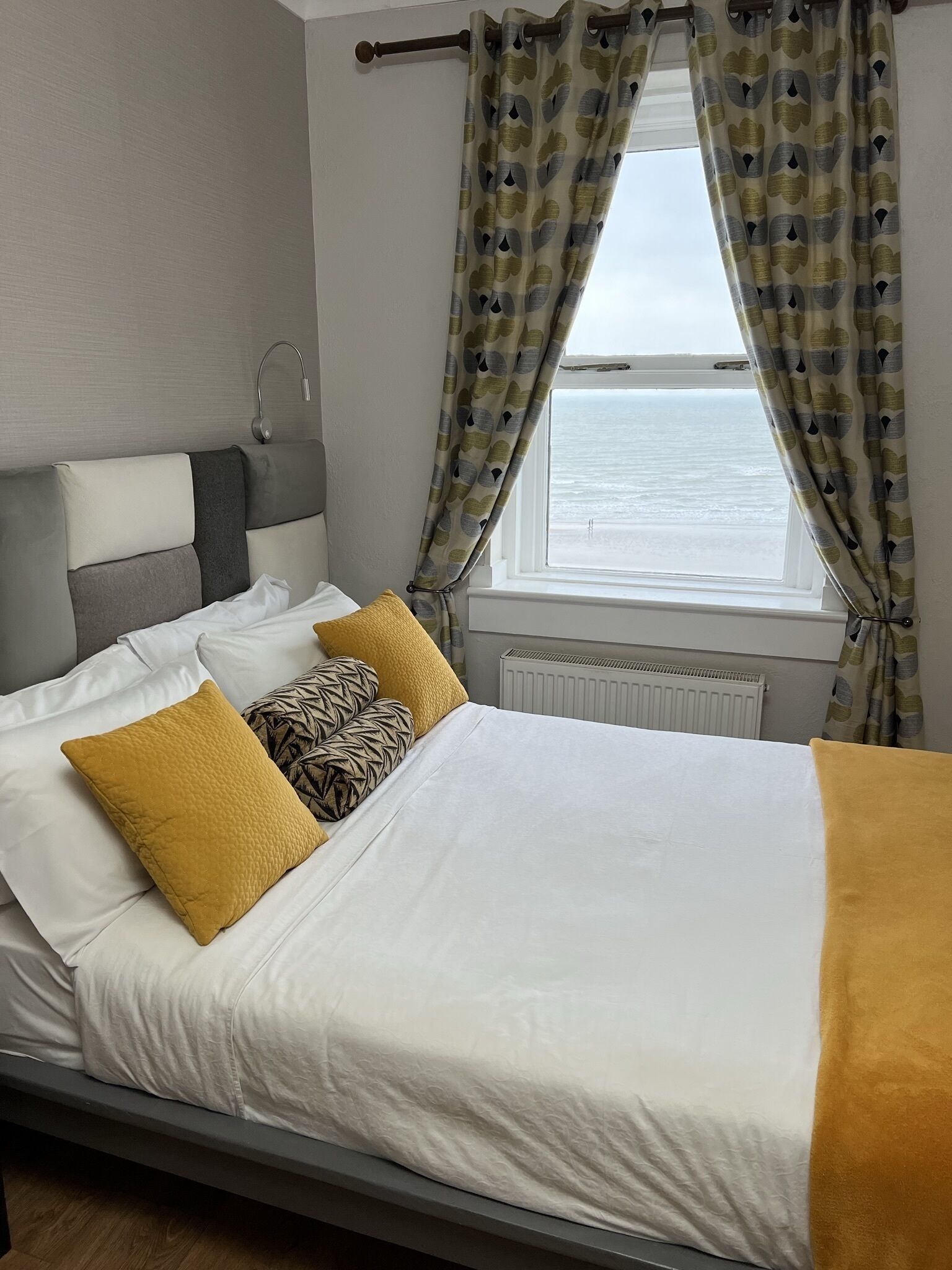 Un letto con lenzuola bianche e gialle senape, due cuscini e vista sul mare attraverso una finestra con tende fantasia.