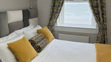 Un letto con lenzuola bianche e gialle senape, due cuscini e vista sul mare attraverso una finestra con tende fantasia.