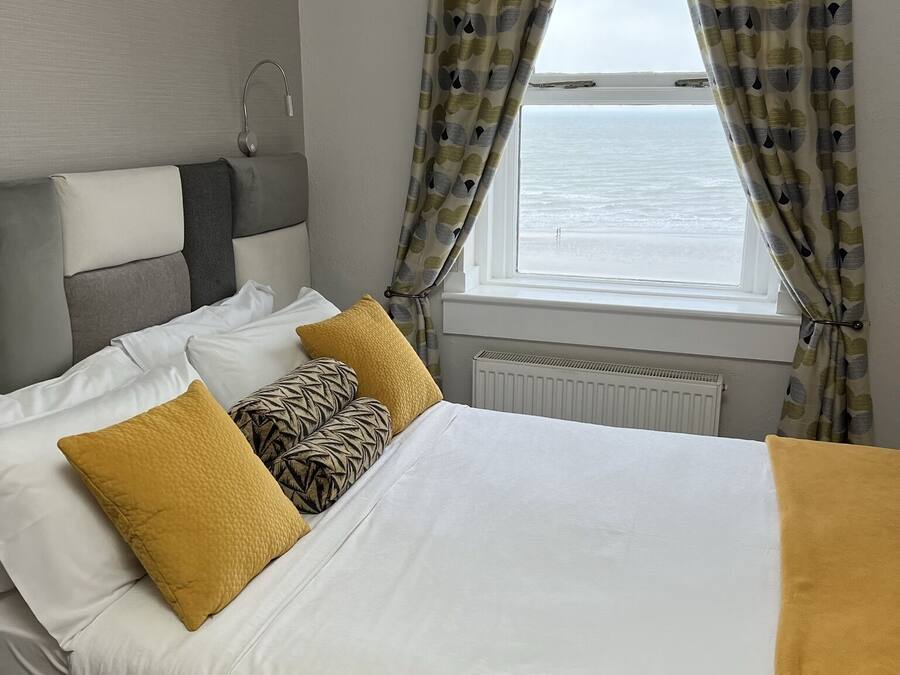 Un letto con lenzuola bianche e gialle senape, due cuscini e vista sul mare attraverso una finestra con tende fantasia.
