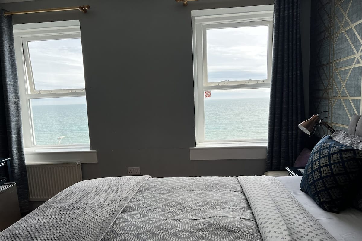 A bedroom with a bed, two windows with a view of the sea, a radiator, and a lamp on the bedside table.