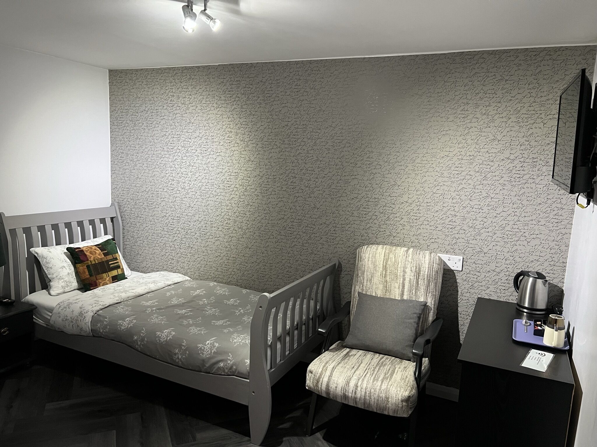 A bedroom with a bed, a chair, a nightstand, a desk, and a kettle.