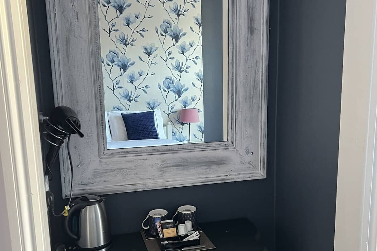A small room with a dark blue wall, a mirror with floral wallpaper, a small table with a kettle, cups, and a tray, and a chair with a patterned cushion.