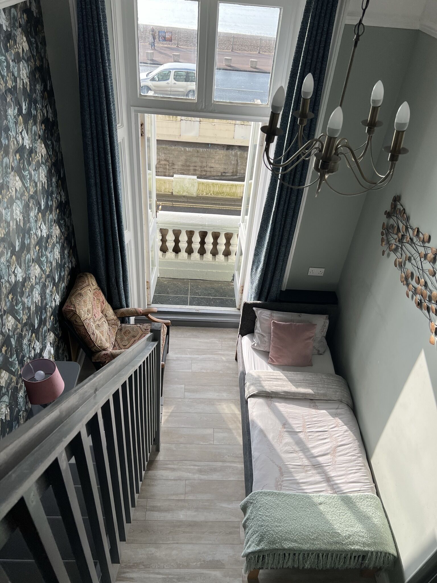 A narrow room with a bed, a chair, and a view of the street through a window.