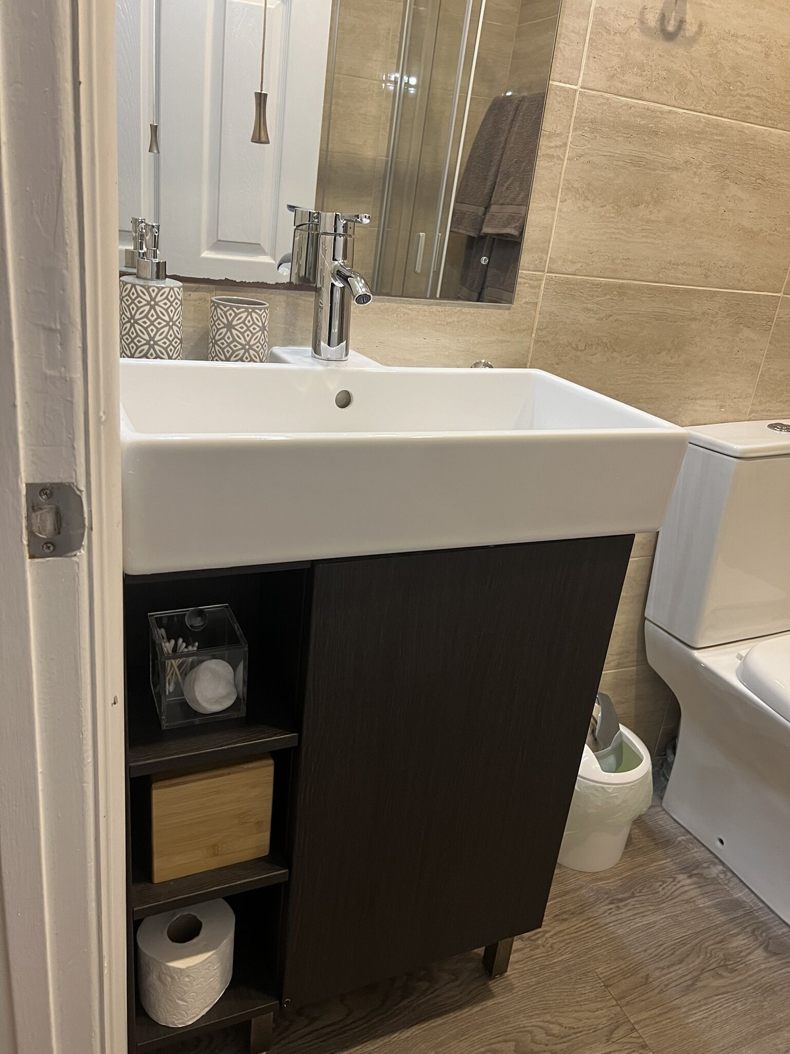 Et badeværelse med en hvid vask, et spejl og et toilet. Der er mønstrede kopper på vasken og en skraldespand ved siden af toilettet.