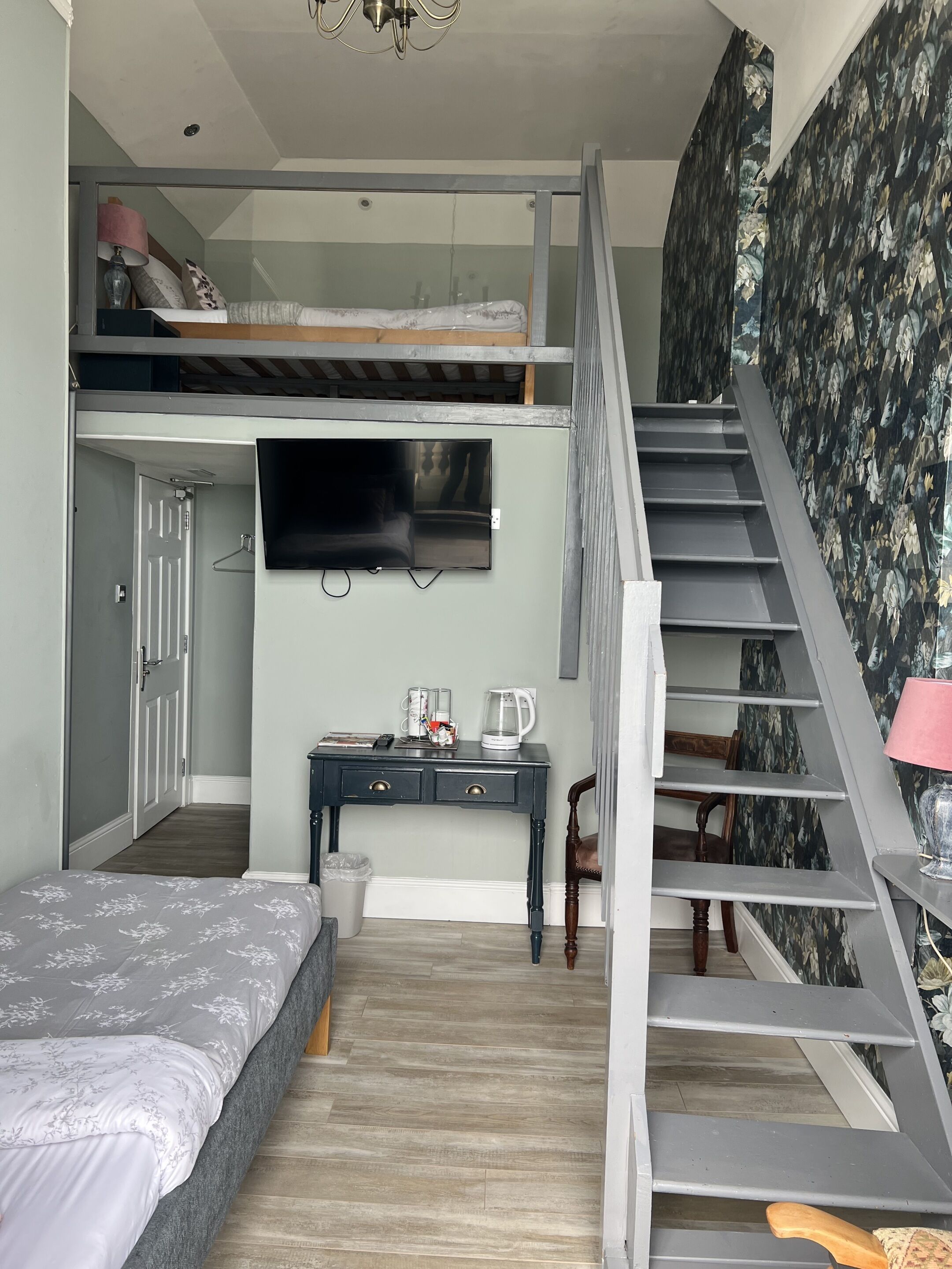 A modern bedroom with a loft bed, a flat-screen TV, a desk with a coffee maker, and a staircase with floral wallpaper.
