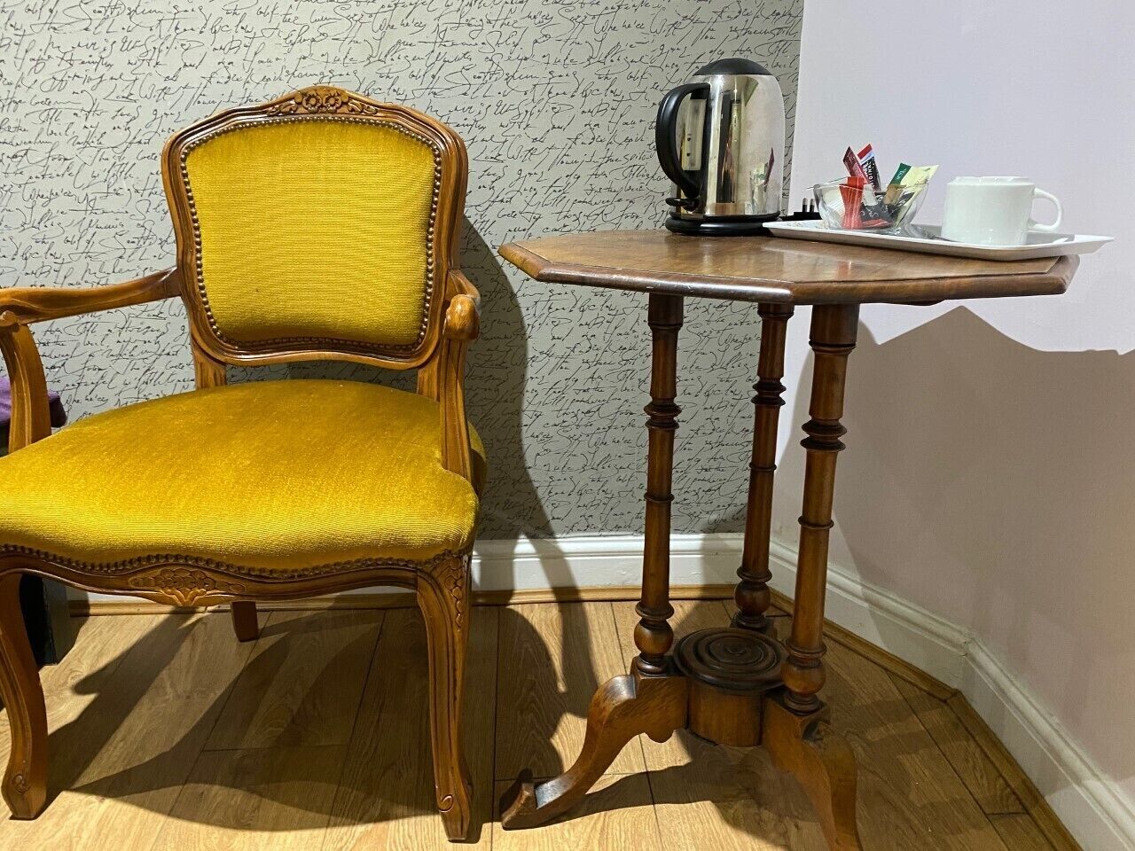 Una butaca amarilla tapizada junto a una mesa de madera con un hervidor y tazas.