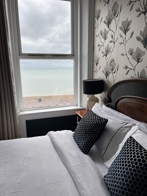 A hotel room with a bed, a nightstand, a lamp, and a view of the sea through a large window.