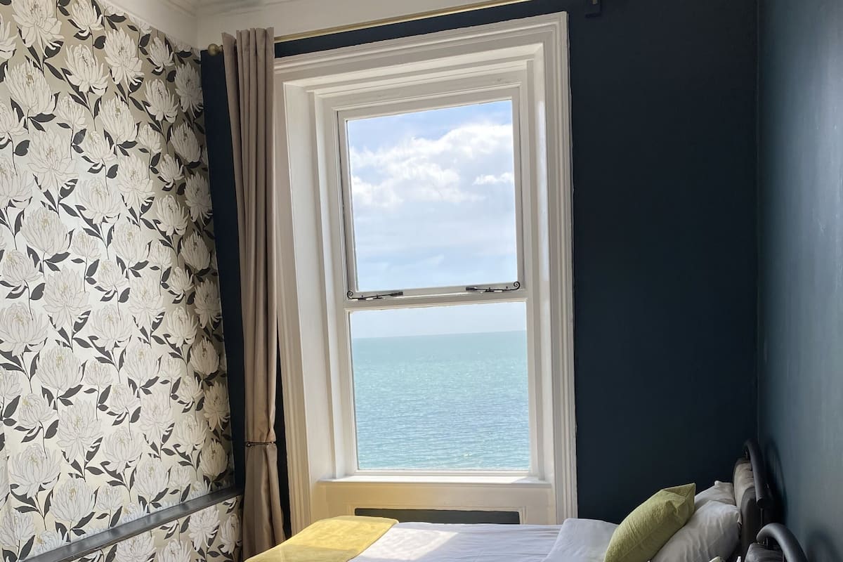 A room with two beds, a window with a view of the sea, and floral wallpaper.