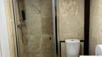 Chuveiro com porta de vidro, vaso sanitário e pia em banheiro com azulejos bege.