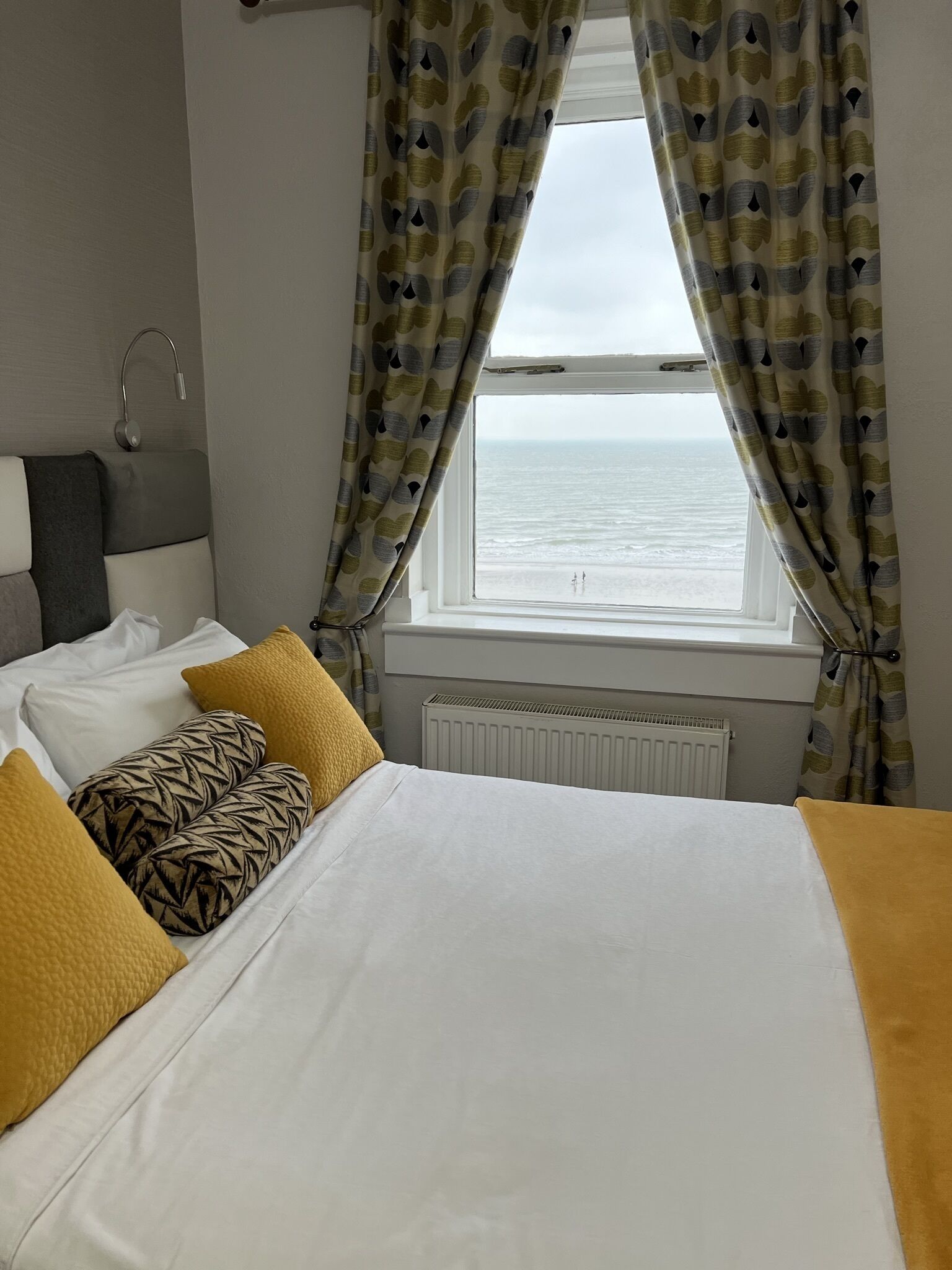 A hotel room with a bed, a window with curtains, a radiator, and a view of the sea.