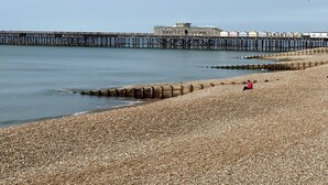 A pebbled beach with a wooden jetty extending into the water, a small building with flags, and a person in red sitting on the sand. - Astral Lodge (Hastings)