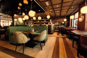 Un espace bar avec un coin salon vert, des tables en bois et des lampes sphériques suspendues.