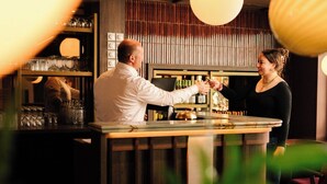 Un espace bar avec un barman et un client. On peut voir des étagères remplies de bouteilles et de verres derrière le bar.