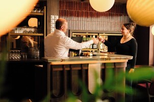 Zona de bar con un cantinero y un cliente. Hay estantes con botellas y vasos detrás de la barra.