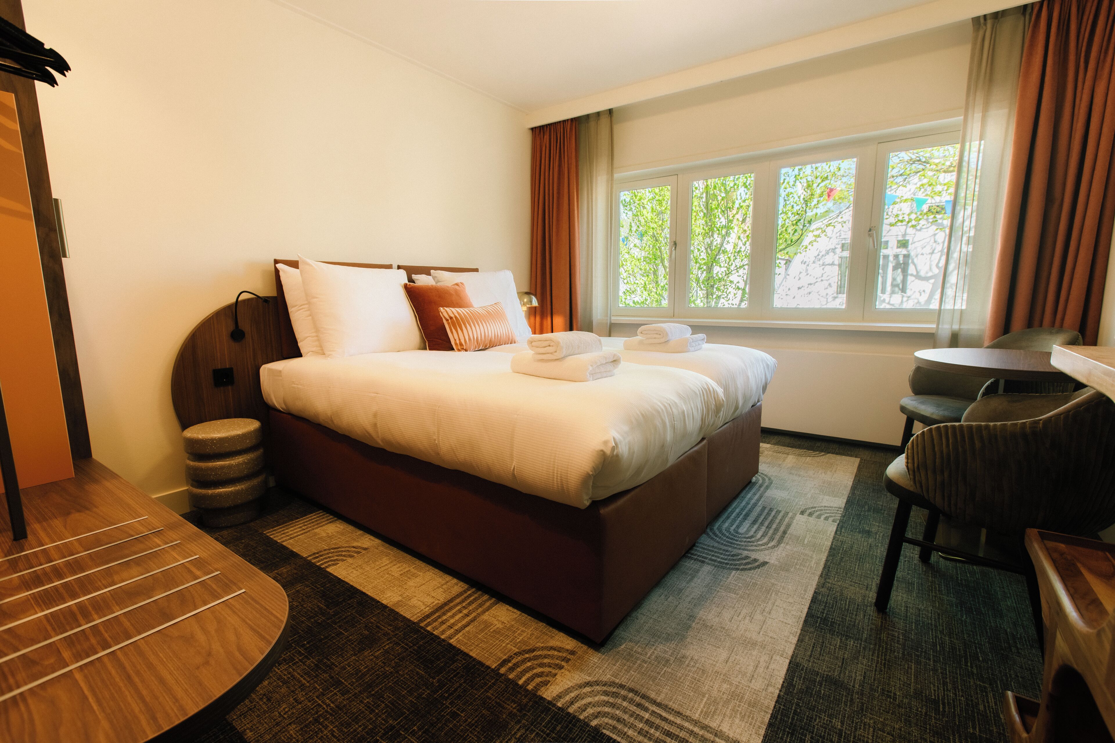 A hotel room with a large bed, a desk, a chair, a small stool, and a window with curtains.