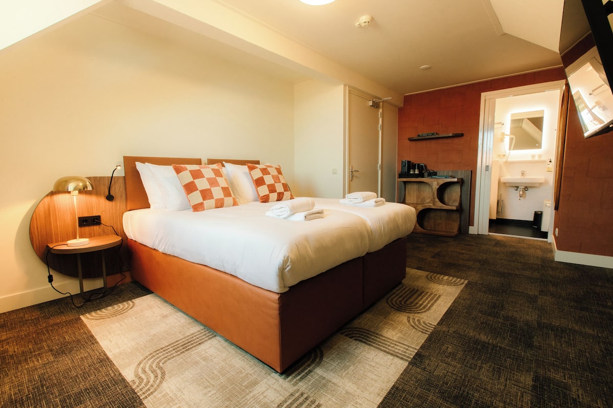 Une chambre d’hôtel avec un lit, une table de chevet, une lampe et une salle de bain visible à travers une porte ouverte.