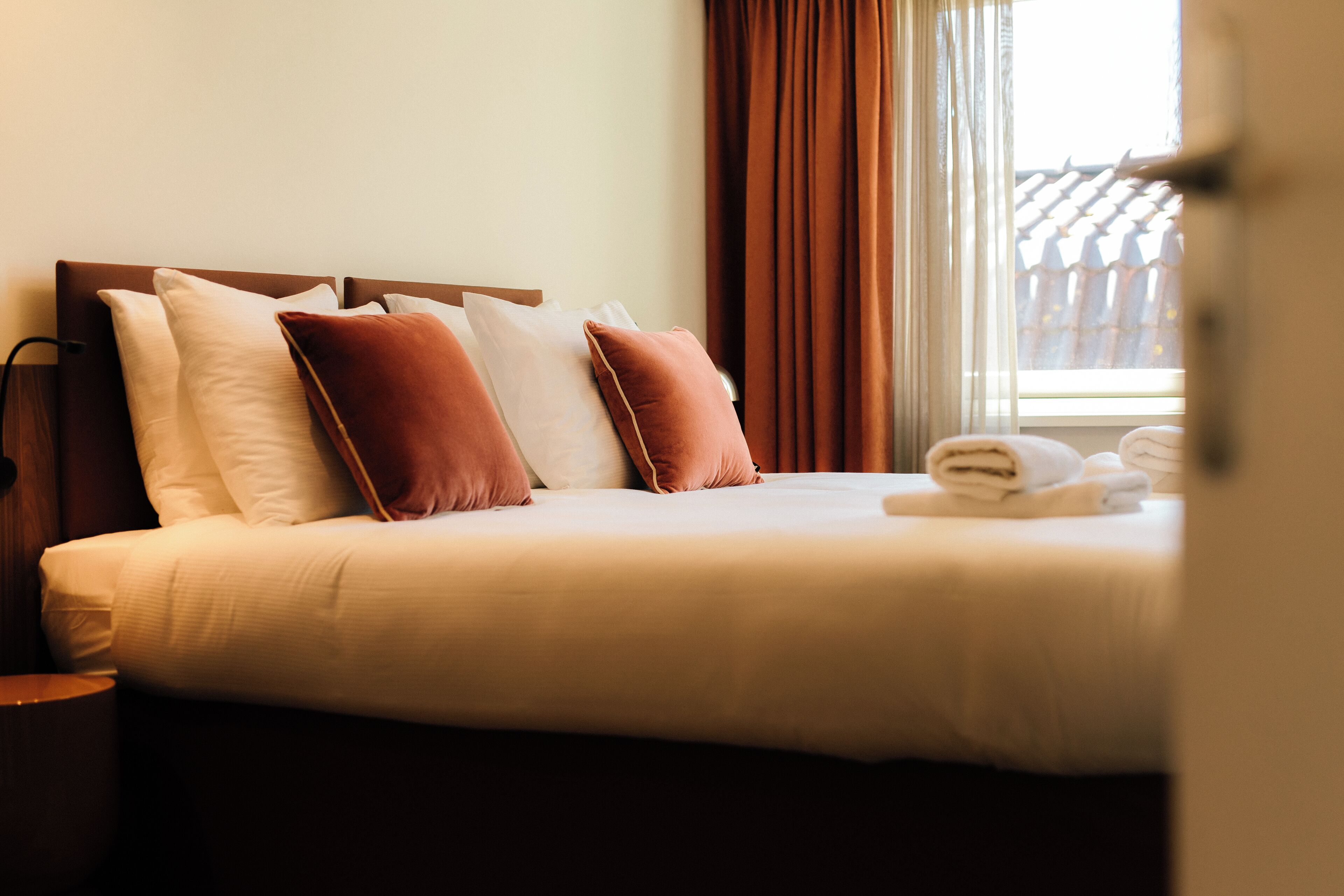 A neatly made bed with pillows, a nightstand, and a window with curtains.