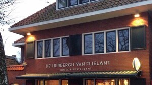 Een gebouw van baksteen met twee verdiepingen, met een bord dat "DE HERBERGH VAN FLIELANT HOTEL RESTAURANT" vermeldt. Voor het gebouw staan buiten tafels en stoelen.