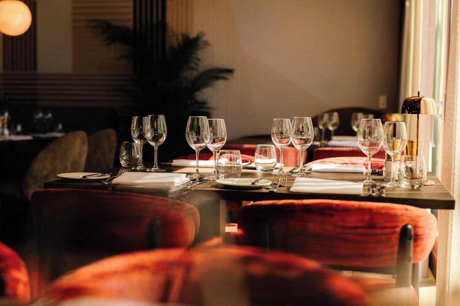 Una mesa de comedor dispuesta con varias copas de vino y platos, sillas tapizadas en rojo y una planta al fondo.