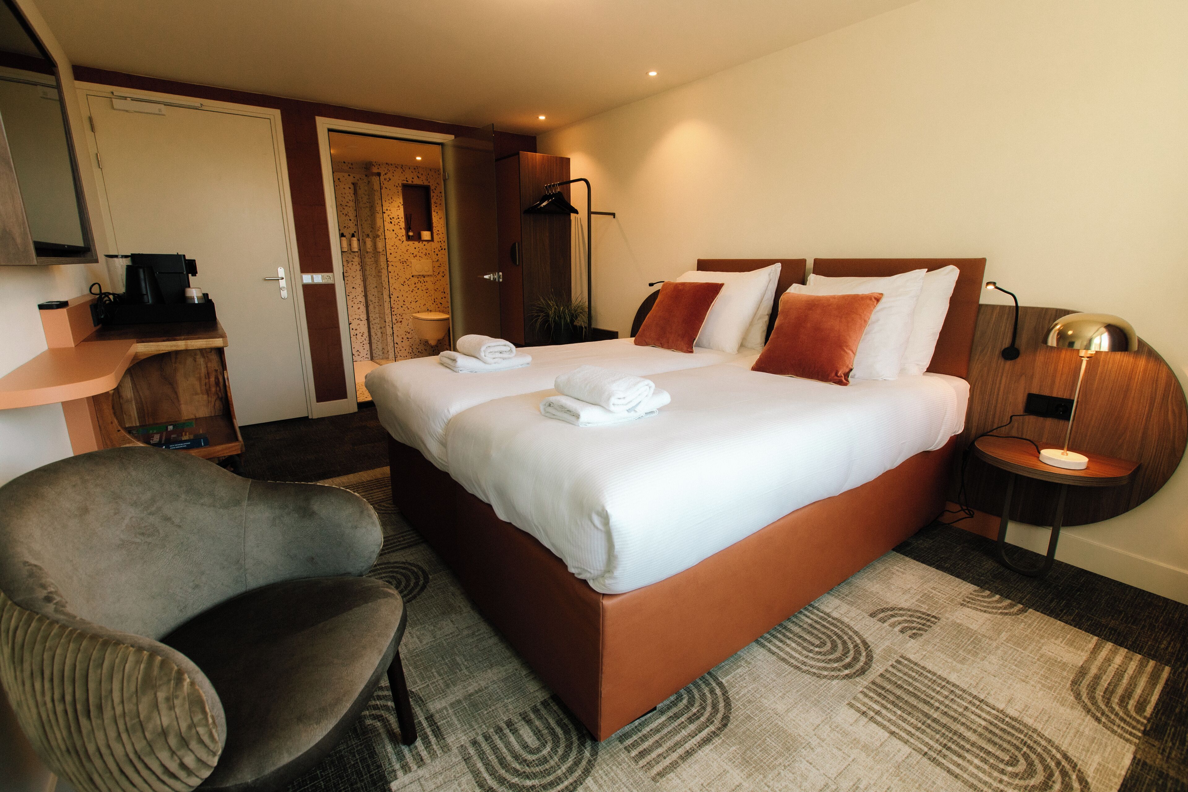A hotel room with a bed, a chair, a desk with a coffee maker, a lamp, and a shower area.