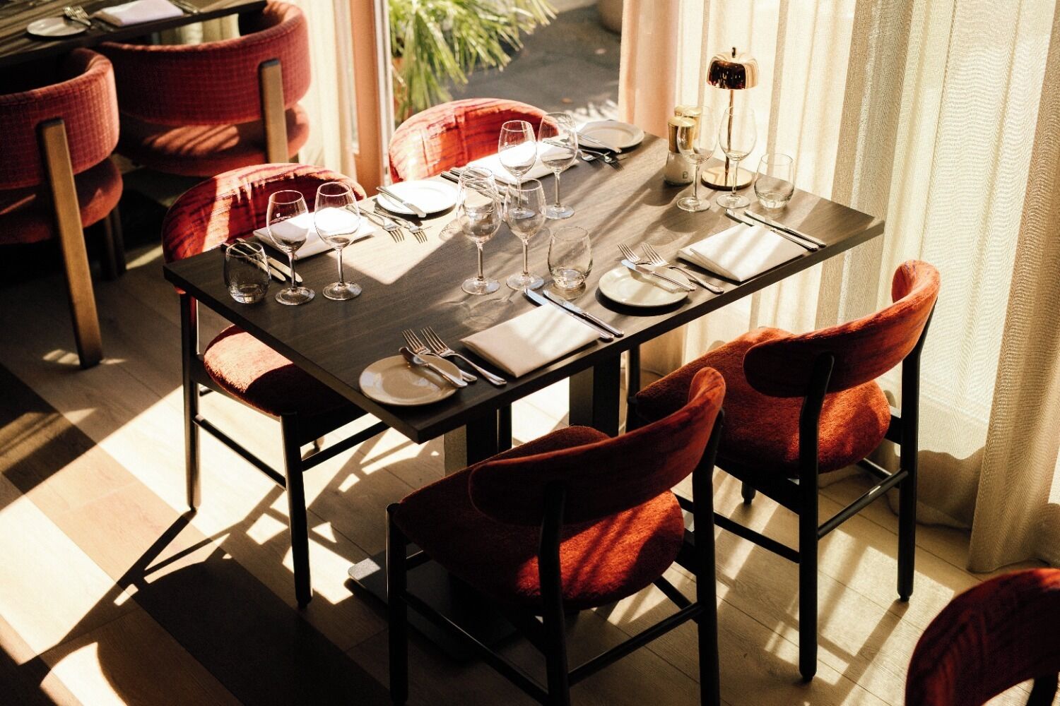 A dining table set with plates, cutlery, and wine glasses, surrounded by red upholstered chairs.