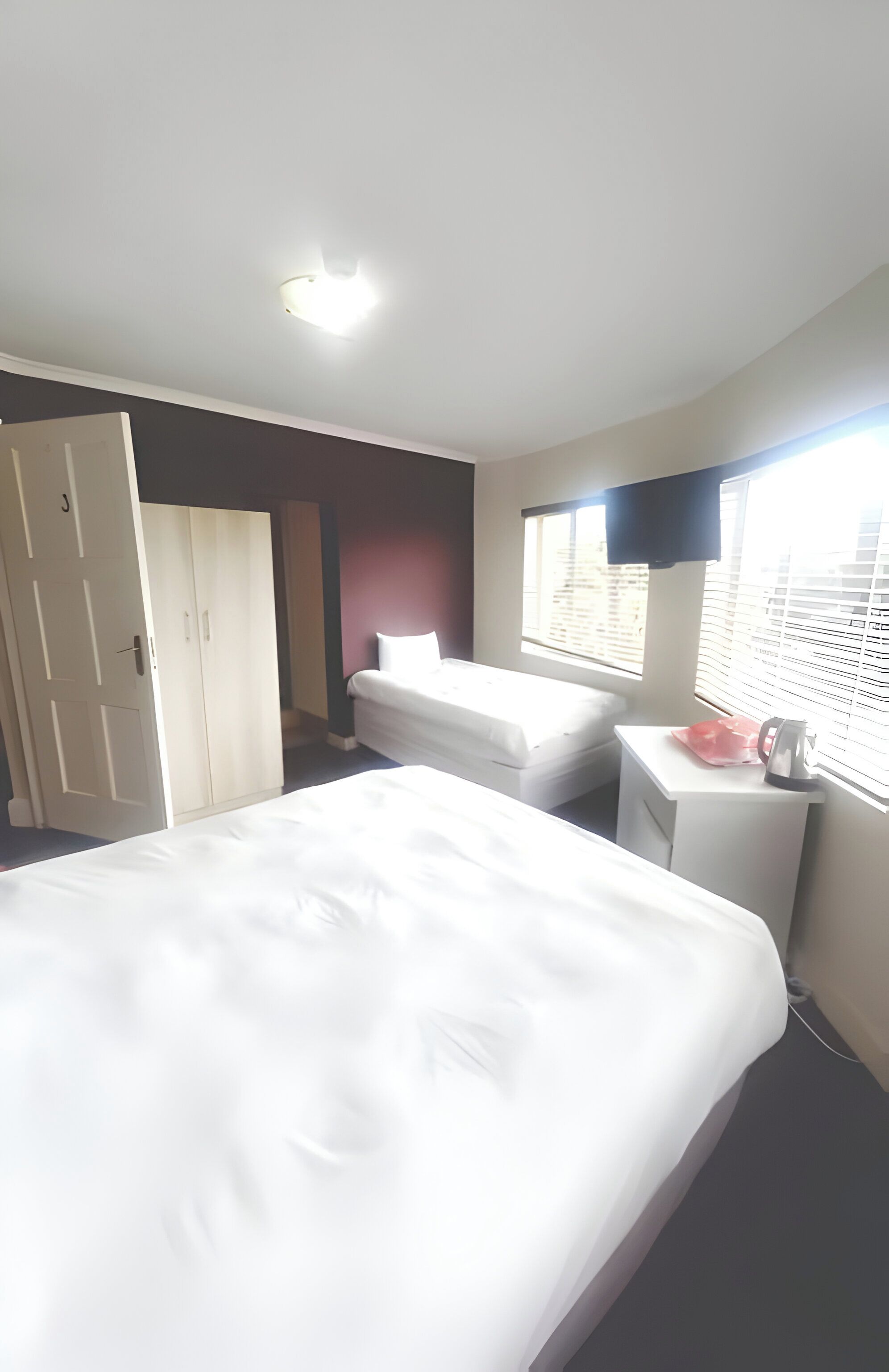 A hotel room with a large bed, a bedside table with a kettle, a wardrobe, and a window with blinds.
