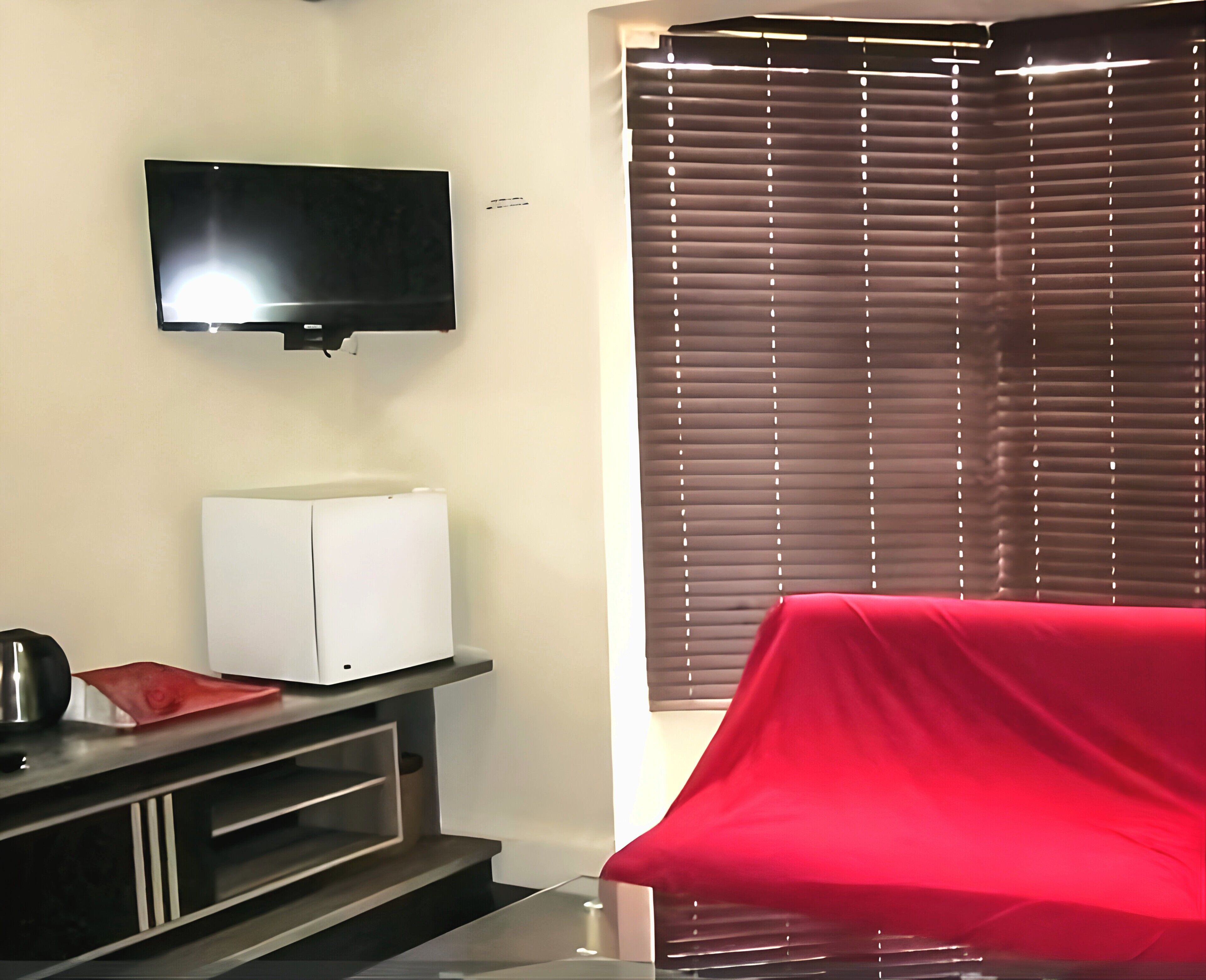 A room with a flat-screen TV mounted on the wall, a white refrigerator, a red-covered object on the floor, and a window with blinds.