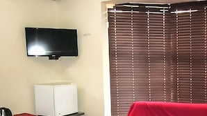 Una habitación con una televisión de pantalla plana montada en la pared, un refrigerador blanco, un objeto cubierto de rojo en el suelo y una ventana con persianas.