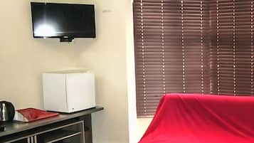 A room with a flat-screen TV mounted on the wall, a white refrigerator, a red-covered object on the floor, and a window with blinds.